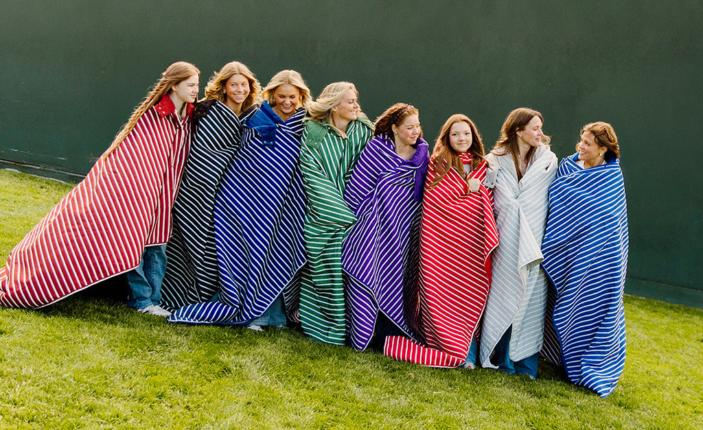A group of women standing together wrapped in different colored striped stadium blankets in a baseball stadium. 