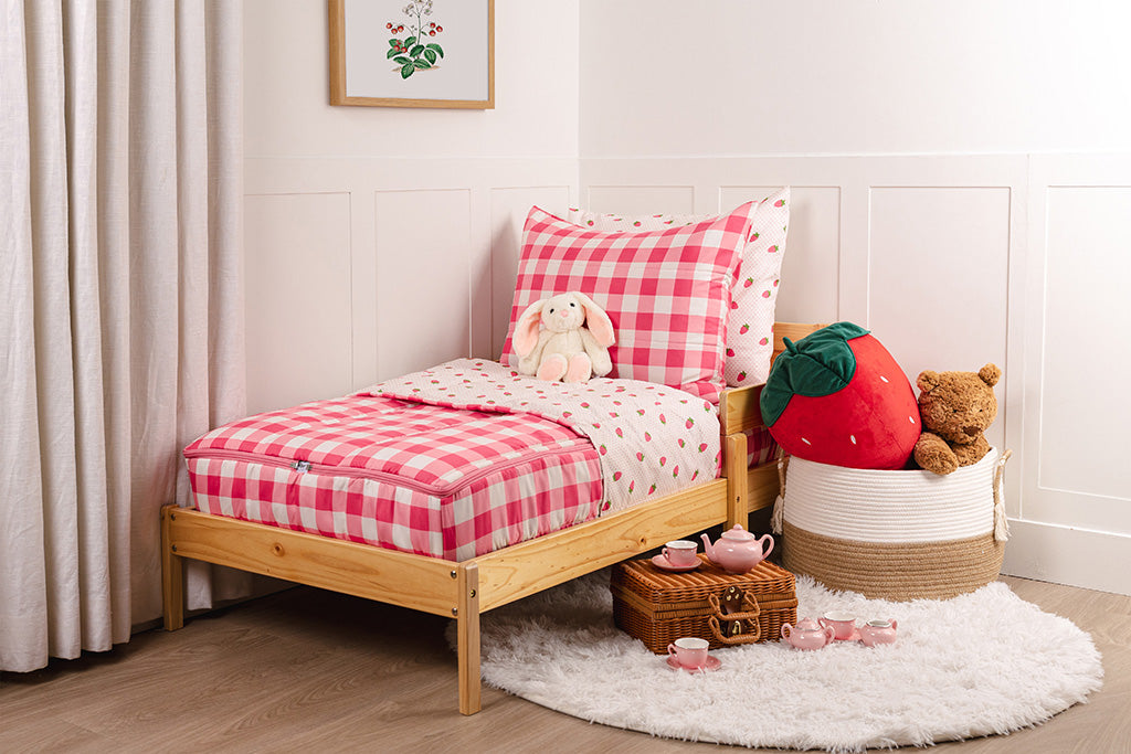 A wooden toddler bed made with pink gingham and strawberry printed zipper bedding and a stuffed animal leaning against the pillows. 