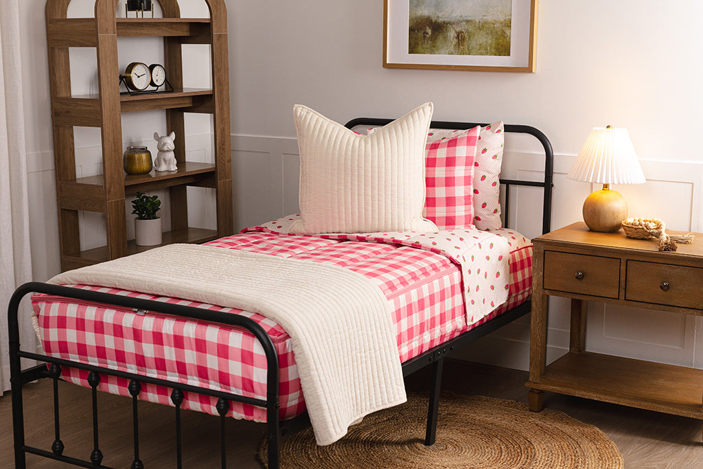 Children zipper bedding in a bedroom on a black bed frame that has a pink gingham exterior and a strawberry print interior, with cream colored accessories. 