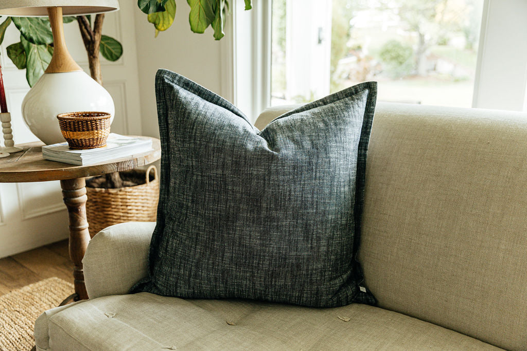 A large charcoal gray pillow arranged on a gray couch by a window and side table.