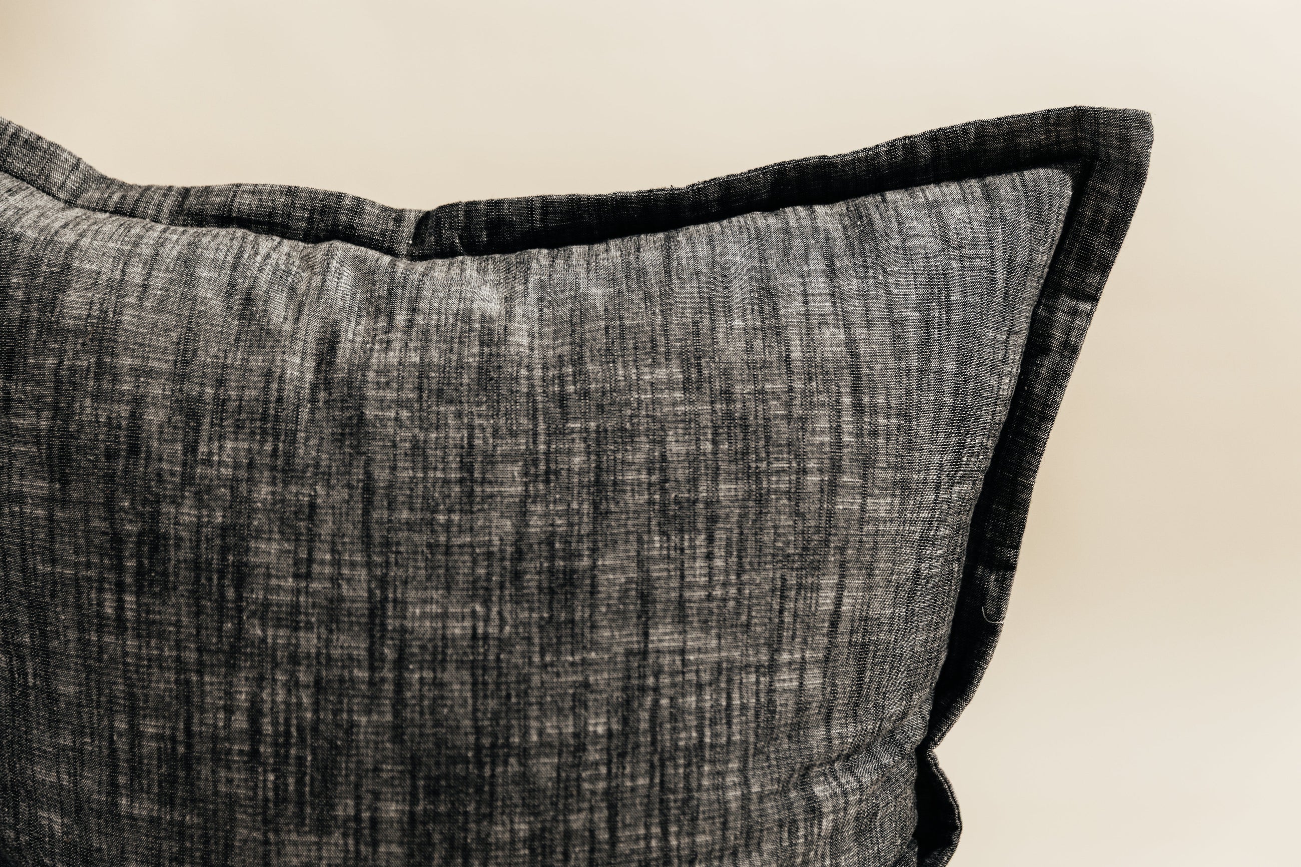 A close up corner view of a large charcoal gray pillow on a cream background.