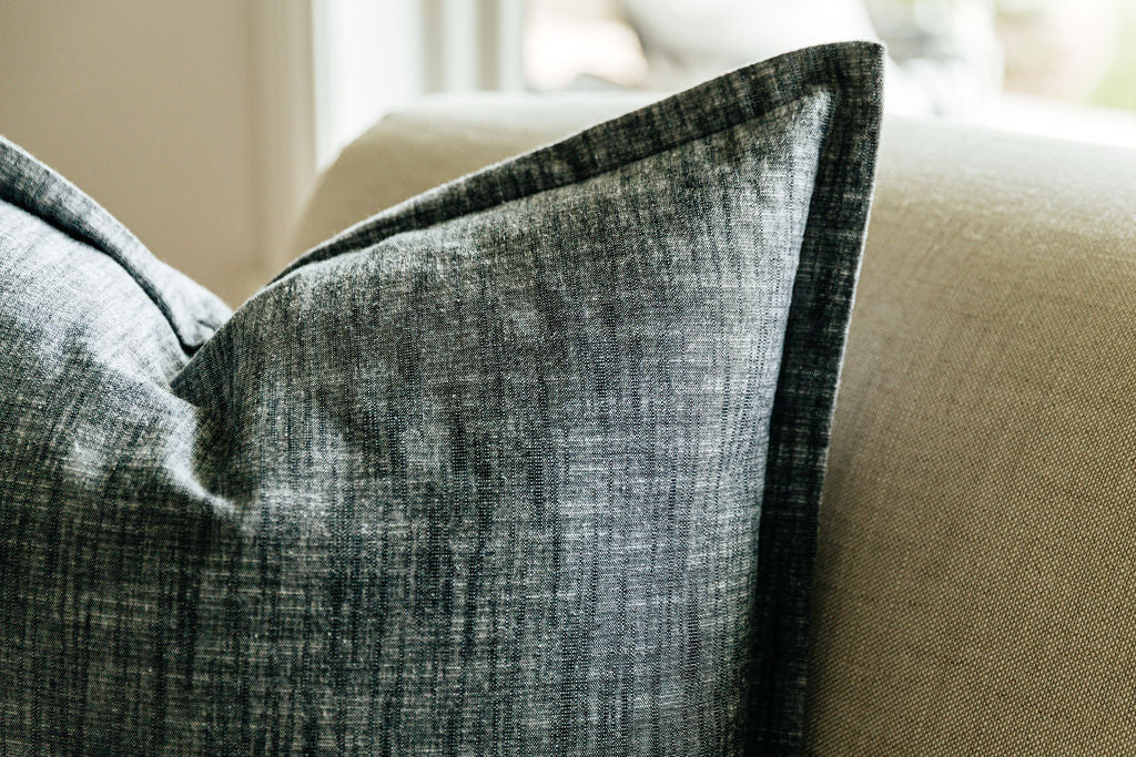 A close up corner view of a large charcoal gray pillow arranged on a gray couch.