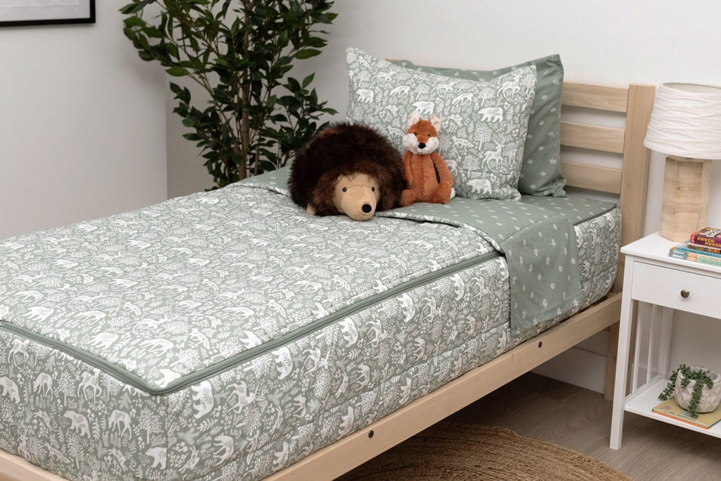 A twin bed made with sage green forest animal kids zipper bedding with a nightstand beside the bed holding books and a lamp.