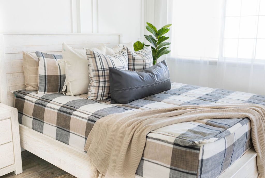 A queen bed made with plaid cream, charcoal gray, and tan zipper bedding and an assortment of neutral pillows and throw blanket.