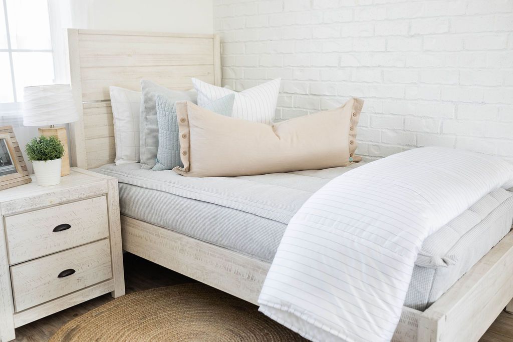 A twin bed made with light gray waffle textured zipper bedding and an assortment of neutral and light blue quilted pillows and white striped blanket.