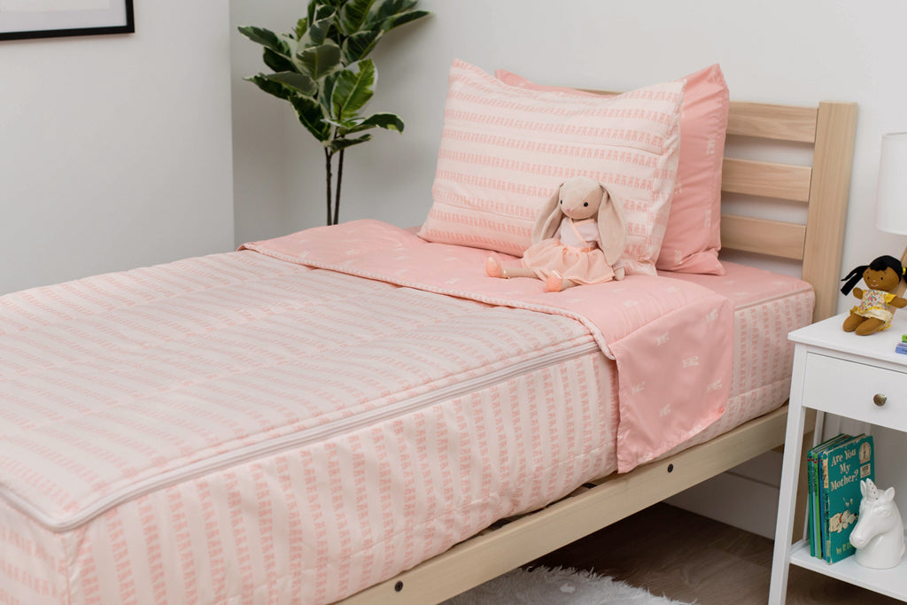 A twin bed made with pink ballet shoes kids zipper bedding, a nightstand beside it with books on the shelf and toys scattered on the floor.