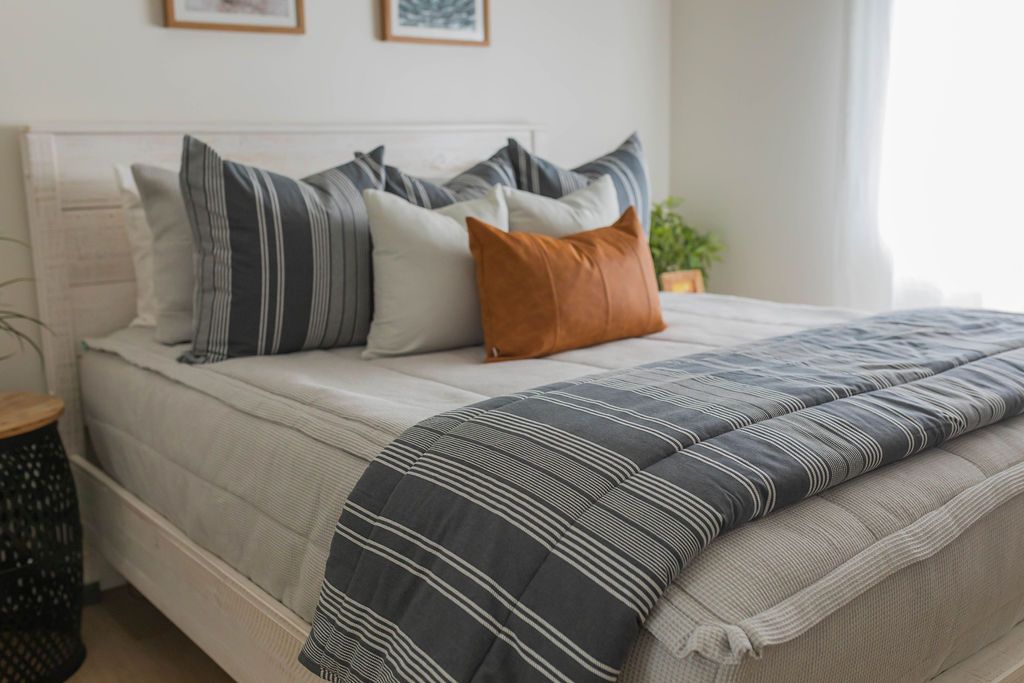 A queen bed made with light gray textured zipper bedding and adorned with neutral and dark gray and white striped pillows and blanket.