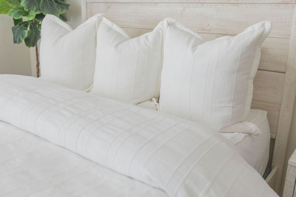A close up view of a bed made with white zipper sheets and a textured white duvet and matching large pillows.