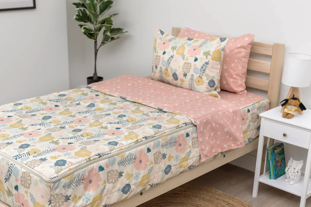 A twin bed made with pink, yellow, and blue floral kids zipper bedding, a nightstand beside it with books on the shelf and toys scattered on the floor.