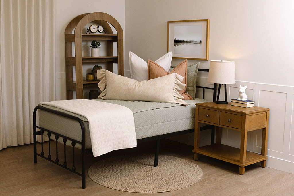 A twin bed made with sage green zipper bedding and neutral and light pink pillows and a floral pattern blanket.