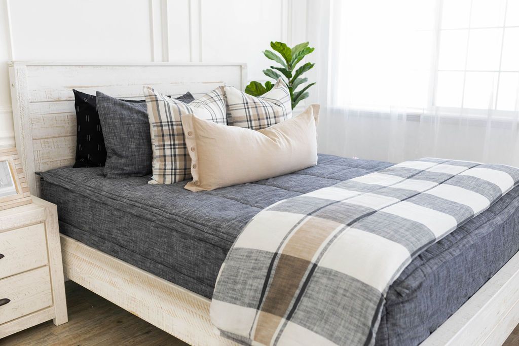 A brightly lit bedroom with a bed made with deep gray zipper bedding and an assortment of neutral pillows and blanket.