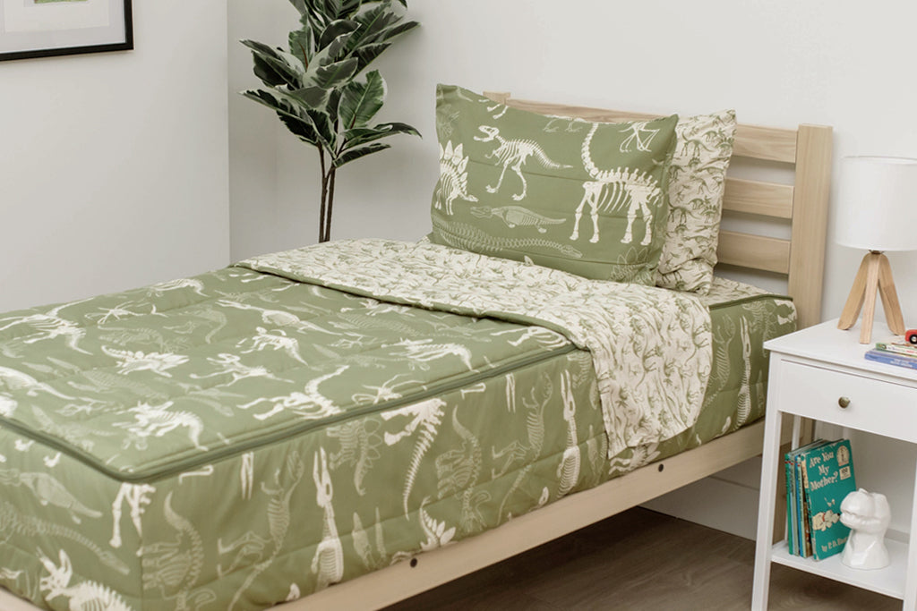 A twin bed made with olive green dinosaur fossils kids zipper bedding, a nightstand beside it with books on the shelf and toys scattered on the floor.