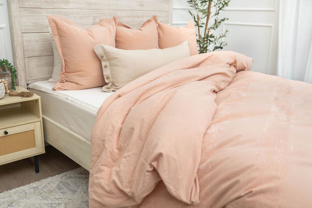 A brightly lit bedroom with a bed made with white zipper sheets and a peach zipper duvet partially folded half way down the bed and adorned with neutral and matching pillows.