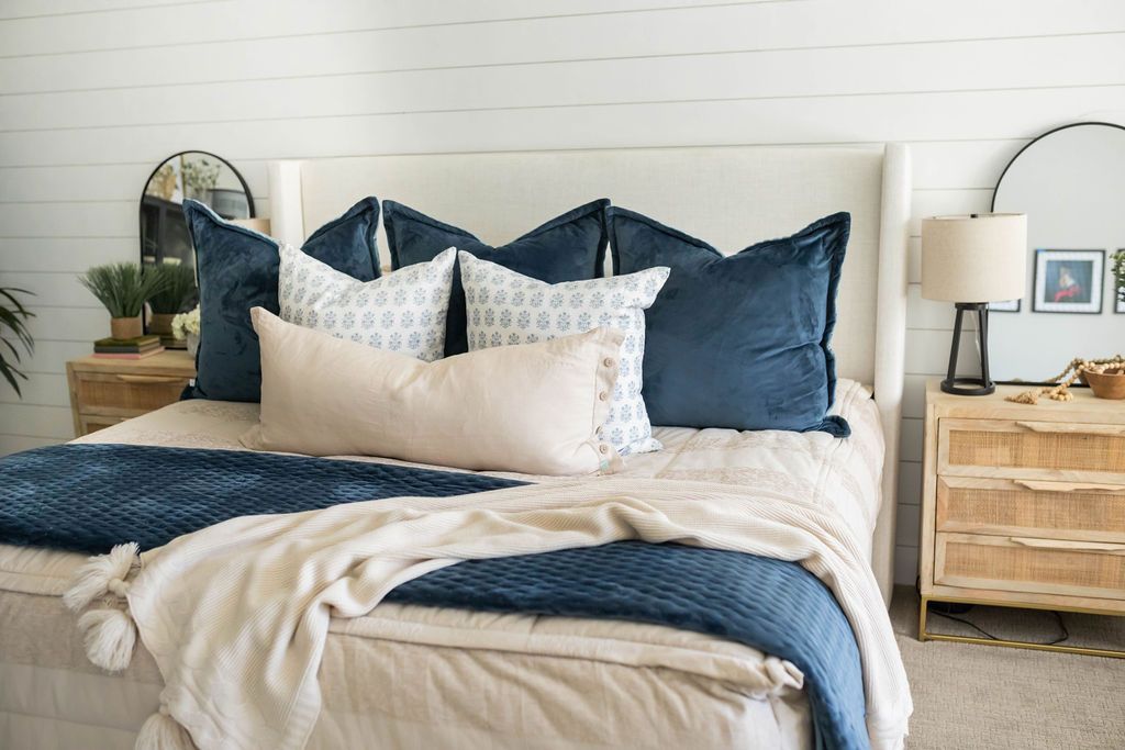 A king bed with a large bed frame made with tan faded stripes zipper bedding and an assortment of neutral and blue velvet pattern throw pillows and matching blankets.