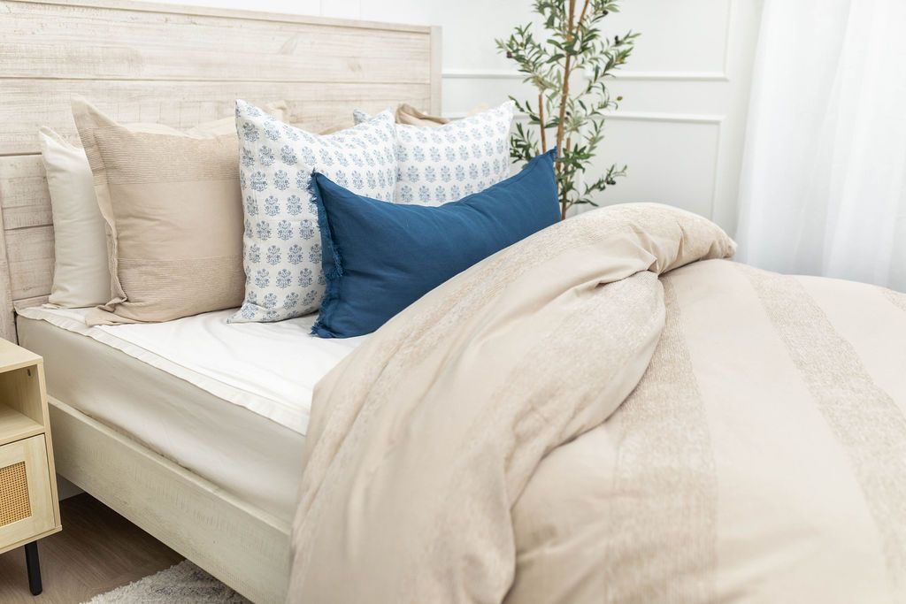 A brightly lit bedroom with a bed made with white zipper sheets and a tan zipper duvet partially folded half way down the bed and adorned with neutral and blue pillows.