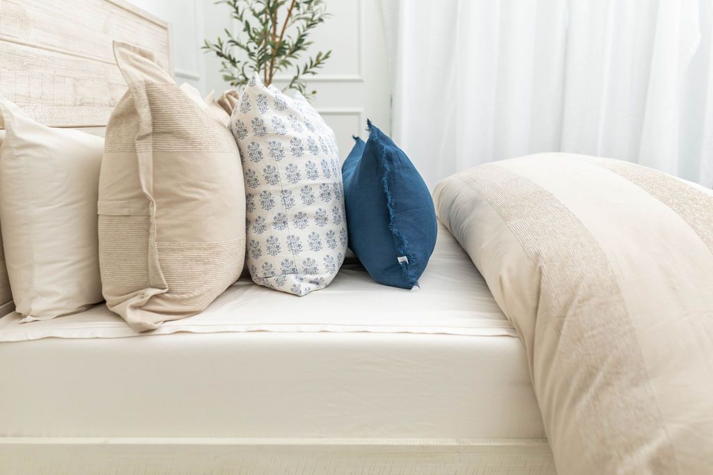 A side view of a bed made with white zipper sheets and a tan zipper duvet folded half way down the bed and adorned with neutral and blue pillows.