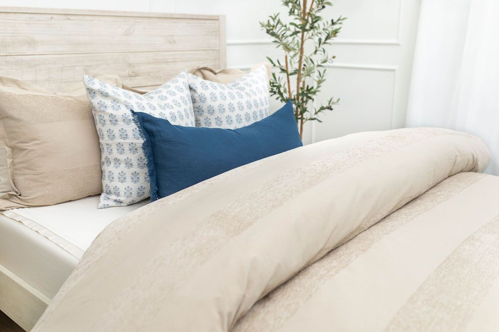 A brightly lit bedroom with a bed made with white zipper sheets and a tan zipper duvet folded half way down the bed and adorned with neutral and blue pillows.