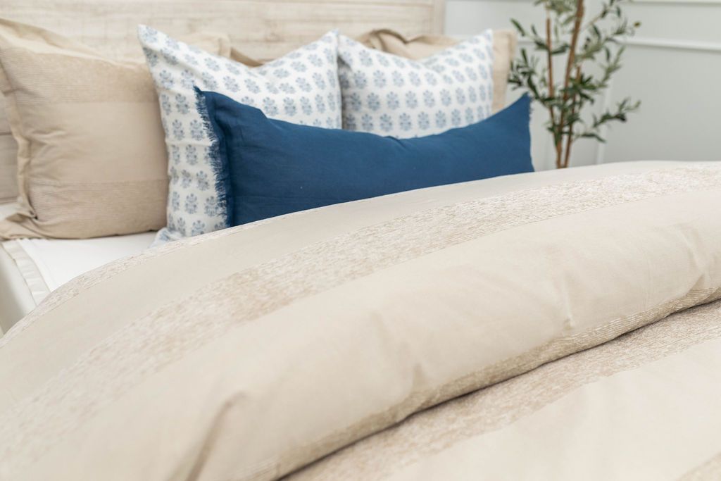 A close up view of a bed made with white zipper sheets and a tan zipper duvet folded half way down the bed and adorned with neutral and blue pillows.