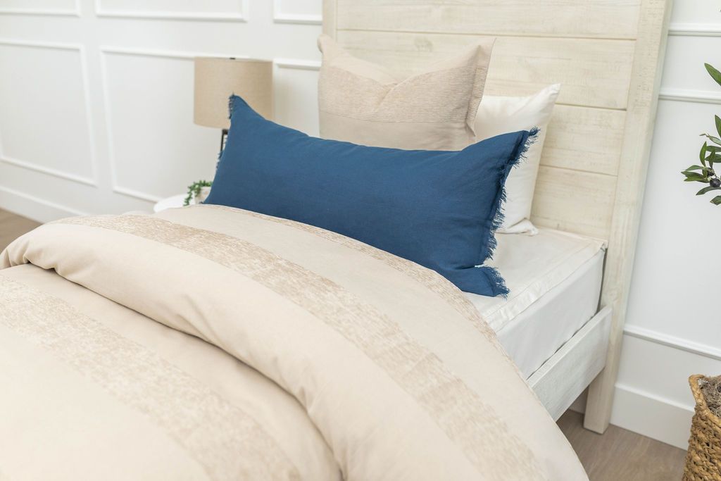 A close up view of a twin bed made with white zipper sheets and a tan zipper duvet folded half way down the bed and adorned with neutral and blue pillows.