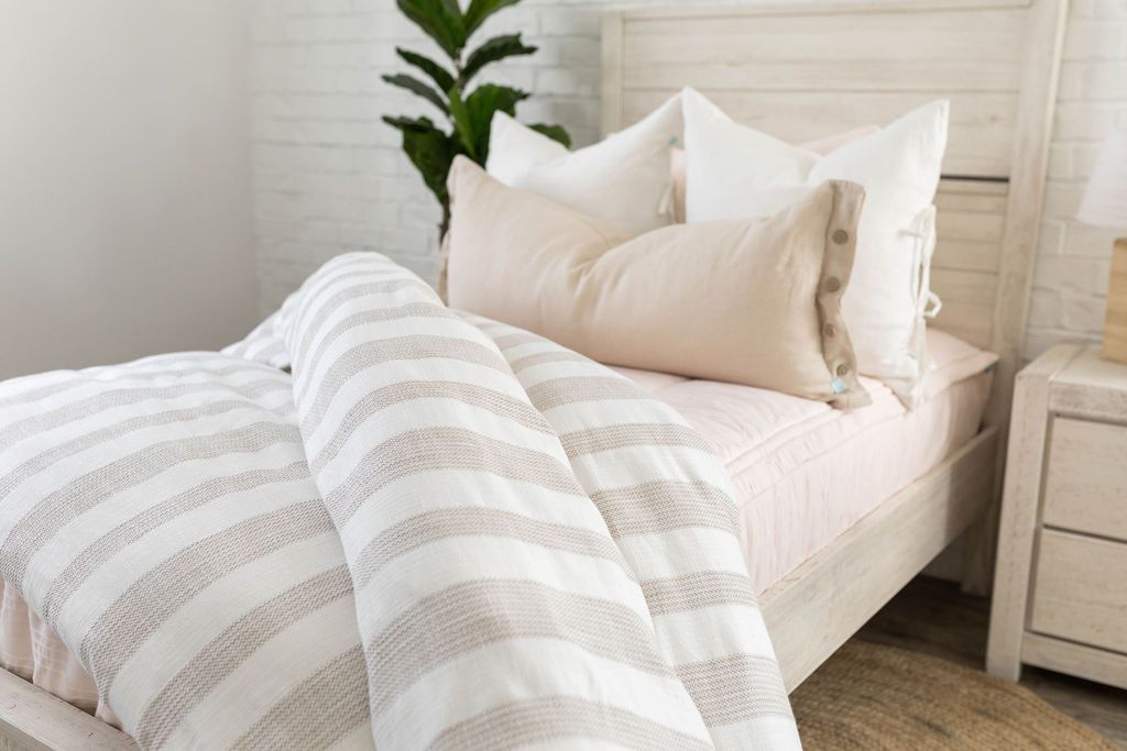 A brightly lit bedroom with a twin bed made with light pink zipper bedding and a woven ivory and tan striped zipper duvet folded partially half way down the bed and adorned with neutral pillows.