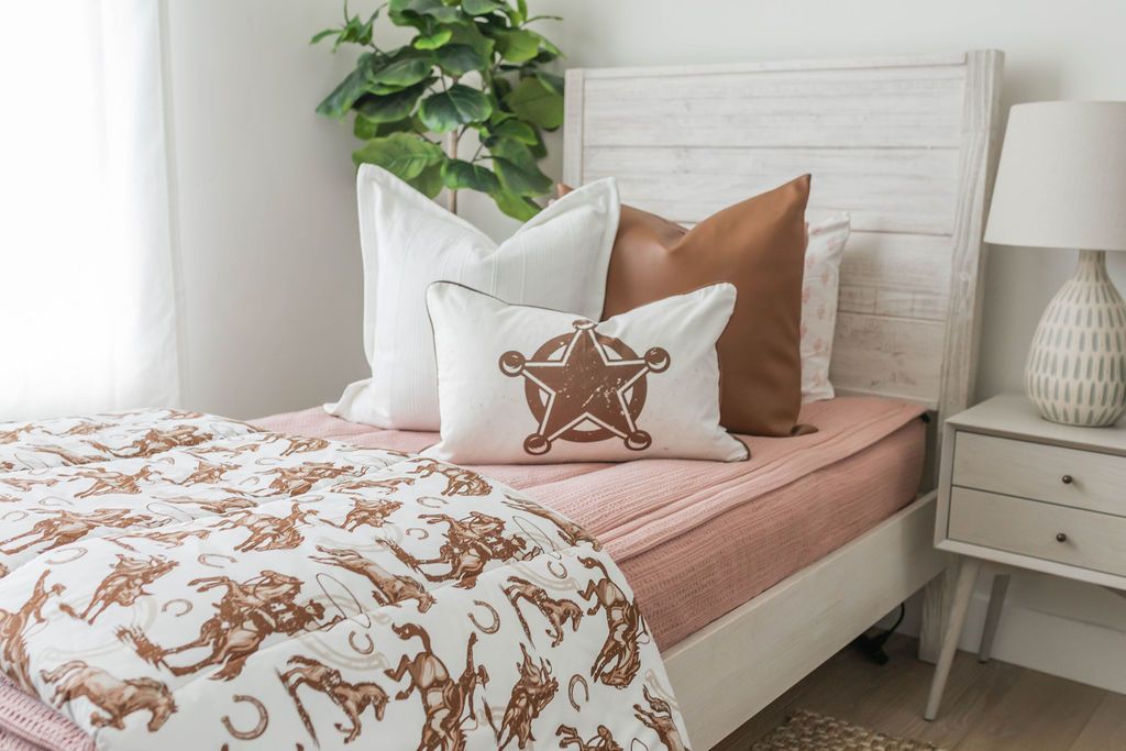 A bed made with pink woven zipper bedding and an assortment of neutral and western throw pillows and matching cowboy printed blanket.