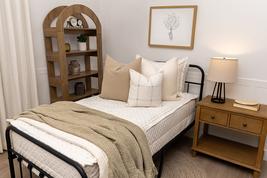 A twin bed made with cream embroidered zipper bedding and adorned with neutral and light pink pillows and a throw tan blanket.