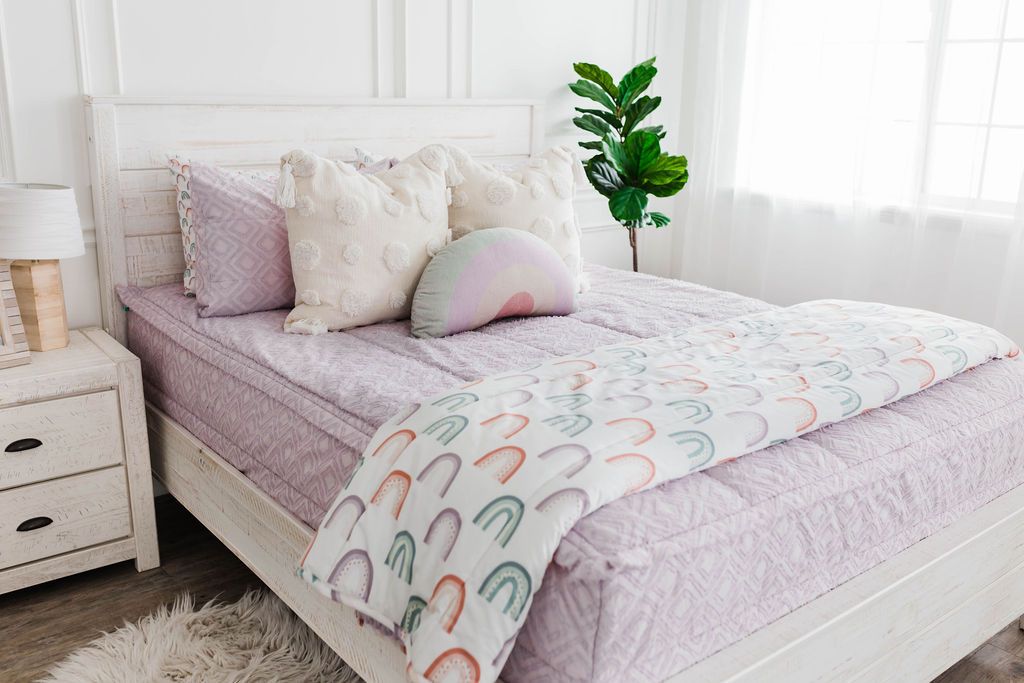 A brightly lit room with a bed made with light purple textured zipper bedding with neutral pillows and a rainbow pattern blanket.