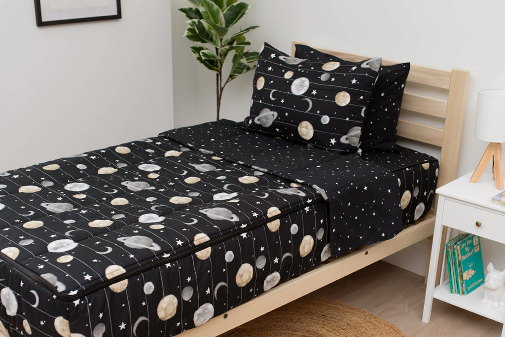 A twin bed made with black planets and stars kids zipper bedding, a nightstand beside it with books on the shelf and toys scattered on the floor.