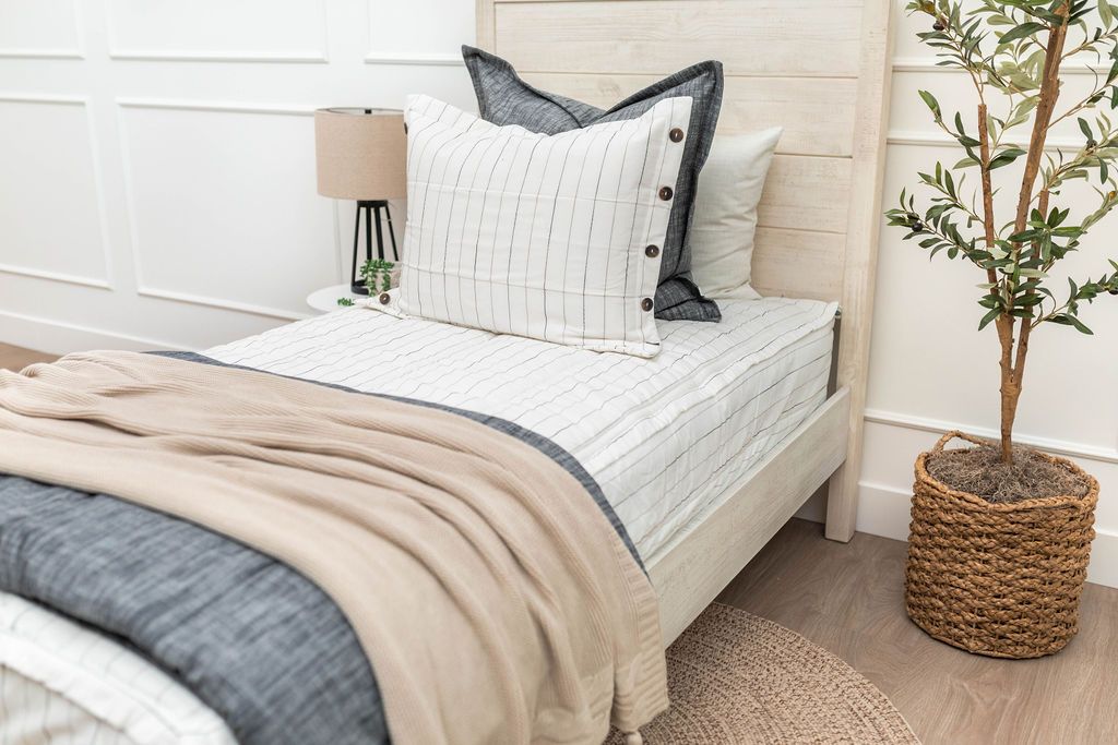 A twin bed made with white and pinstripes zipper bedding and an assortment of neutral and dark gray pillows and blankets.